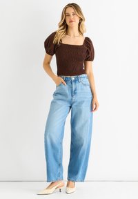 Brauntöniges Smock-Top mit Puffärmeln. Hellblaue, hoch taillierte, weit geschnittene Jeans. Cremefarbene, spitze Slipper. Schlichte Silberkette.