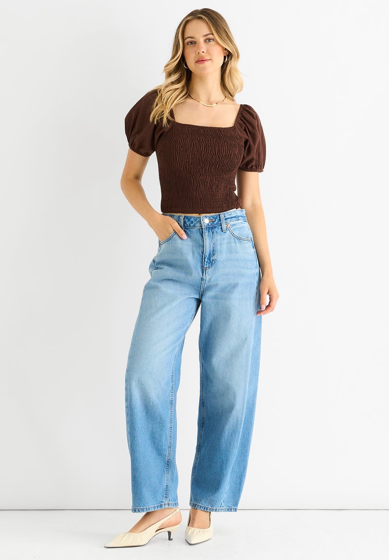 Brauntöniges Smock-Top mit Puffärmeln. Hellblaue, hoch taillierte, weit geschnittene Jeans. Cremefarbene, spitze Slipper. Schlichte Silberkette.