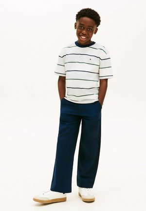 Lächelnder Junge steht mit den Händen in den Taschen einer marineblauen Hose, trägt ein weißes gestreiftes T-Shirt und weiße Turnschuhe vor einem einfarbigen weißen Hintergrund.