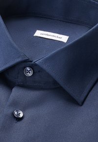 Nærbillede af en mørkeblå skjorte med to knapper knappet og en hvid etiket med teksten "seidensticker" inde i kraven.