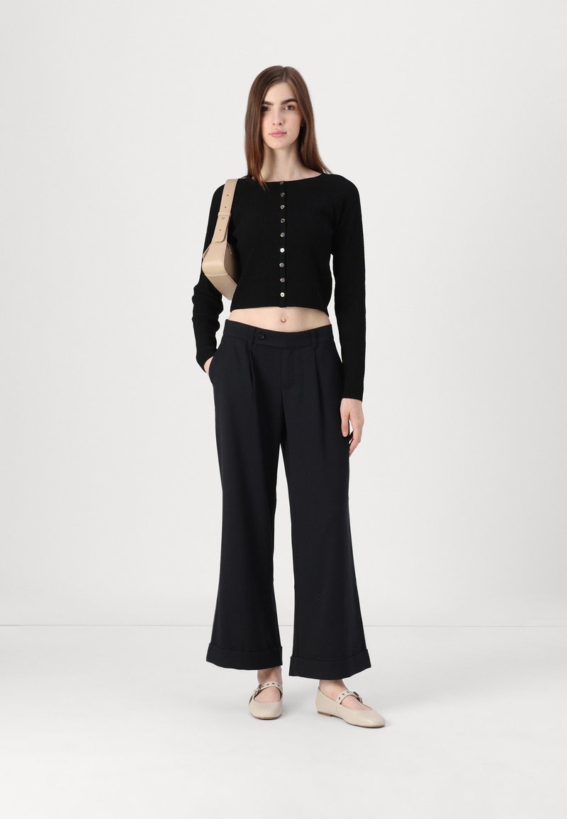 Top corto negro de canalé con detalles de botones, combinado con pantalones negros de corte suelto y zapatos beige de slip-on. Diseño sencillo y colores neutros.