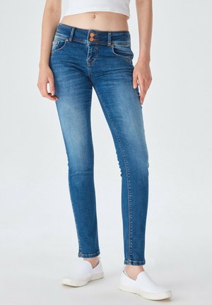 Vrouw draagt vervaagde blauwe skinny jeans met dubbele knoopsluiting en witte instappers, staand tegen een effen achtergrond.