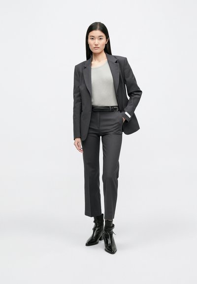 Γκρίζα καμπαρντίνα προσαρμοσμένη πάνω από ένα ανοιχτό γκρι ριγέ τοπ, συνδυασμένη με στενές γκρίζες παντελόνες και μαύρες μποτάκια μέχρι τον αστράγαλο. Απλός σχεδιασμός, δομημένη εφαρμογή.