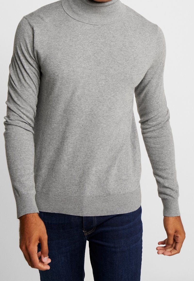 Grauer gestrickter Rollkragenpullover mit langen Ärmeln, gerippten Bündchen und Saum; glatte Textur und tailliertes Design. Getragen über dunklen Jeans.