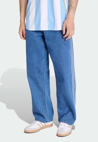 Pantaloni in denim blu a gamba larga con strisce laterali bianche, abbinati a una camicia a righe azzurre e bianche e sneaker bianche con suole beige.