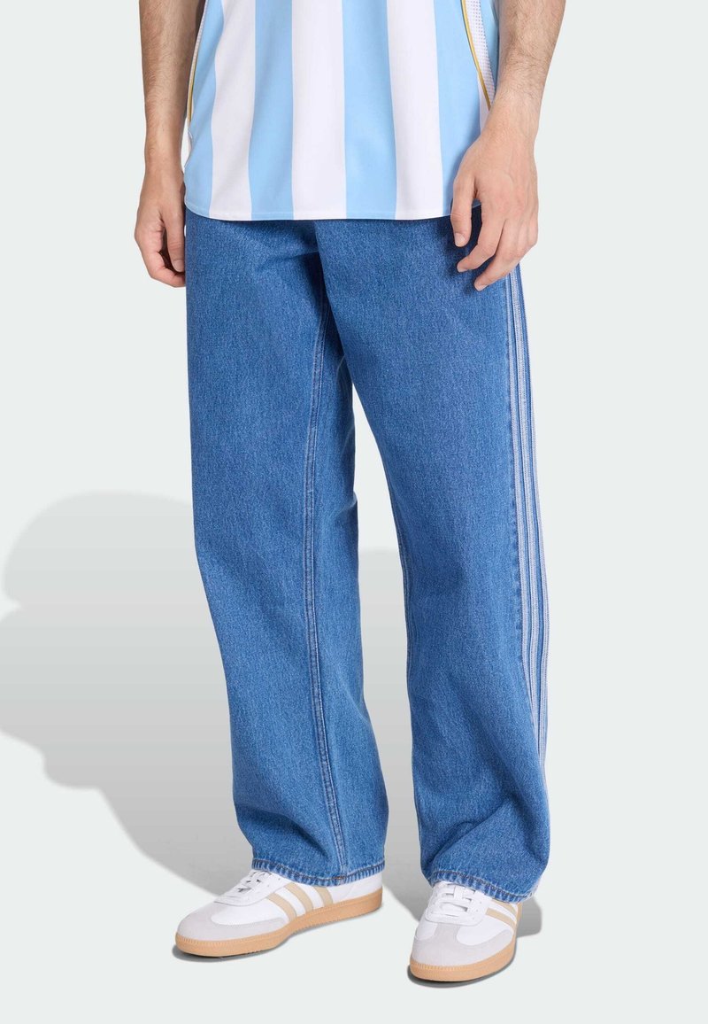 Pantaloni in denim blu a gamba larga con strisce laterali bianche, abbinati a una camicia a righe azzurre e bianche e sneaker bianche con suole beige.
