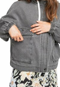Chaqueta de mezclilla gris con puños elásticos, un bolsillo frontal y cordón blanco. Combinada con una falda de estampado floral en la parte inferior.