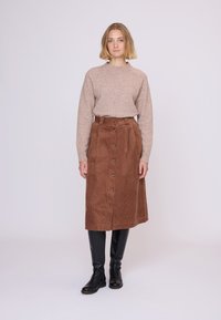 Brun fløjlskørt med lukning foran med knapper, parret med en lys beige sweater. Modellen bærer sorte knæhøje støvler.