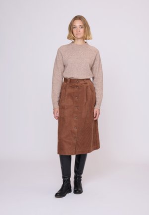 Brun fløjlskørt med lukning foran med knapper, parret med en lys beige sweater. Modellen bærer sorte knæhøje støvler.