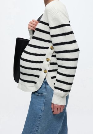 Person iført en hvid sweater med sorte horisontale striber og knapper i siden, kombineret med blå jeans og bærende en sort skuldertaske.