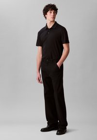 Crna polo majica s kratkim rukavima uparena s crnim hlačama i crnim cipelama. Tkanina izgleda glatko i krojena za prilagođeni izgled.