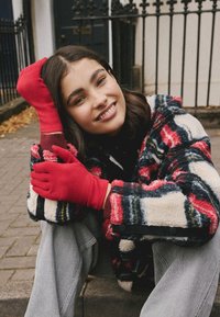 Veste en polaire avec un motif audacieux rouge, bleu marine et crème ; associée à des gants rouge vif et un jean gris, sur un fond extérieur texturé.