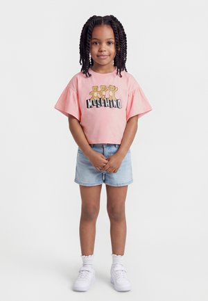 Mladé dievča s vrkočmi, oblečené v ružovom tričku s potlačou medvedíka, riflových kraťasoch, bielych ponožkách a bielych teniskách, stojace so zloženými rukami.