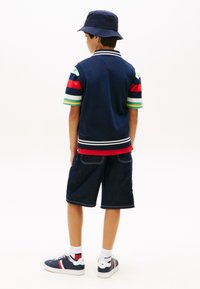Αγόρι που στέκεται πλάτη, φοράει σκούφο μπλε ναυτικού τύπου, ριγέ κοντομάνικο πουκάμισο, μπλε γιλέκο, μακριά σορτς από τζιν, λευκές κάλτσες και αθλητικά παπούτσια.