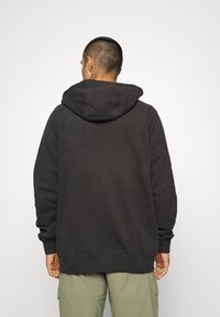 Sweatshirt à capuche noir en matériau doux. Présente une coupe décontractée, des poignets côtelés et une grande poche arrière. Porté avec un pantalon kaki.