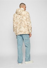 Bēša kamuflāžas kapuci jaka, viegla materiāla, elastīgi manšeti, kapucis ar auklu, kombinēta ar gaiši ziliem platiem džinsiem un baltiem sporta apaviem.