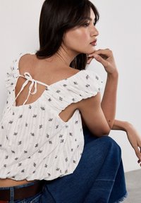 Blusa bianca con texture, ricami floreali, maniche a balza e design con laccio posteriore. Indossata con una gonna di jeans, mostra un dettaglio con schiena scoperta.