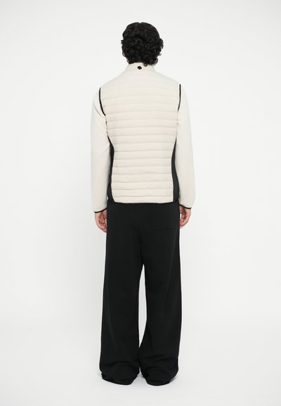 Lys beige polstret vest med høy krage, svarte sidestykker og brede svarte bukser. Strukturelt stoff med en glatt overflate.