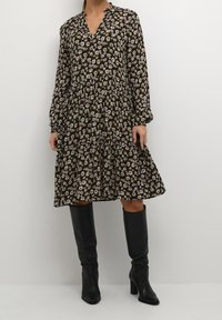 Femme portant une robe florale à manches longues et longueur genou, associée à des bottes à talons en cuir noir jusqu'aux genoux, sur un fond uni.