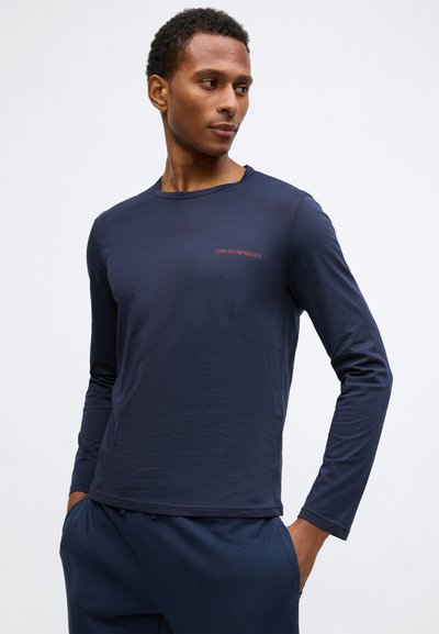Sötétkék, hosszú ujjú póló sima anyagból, apró piros "EMPORIO ARMANI" logóval a mellkason, kerek nyakkivágással és laza szabással.
