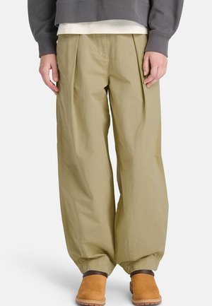 Leveälahkeiset beige-väriset housut, joissa on kerätyt joustavat lahkeet, päällä seisovalla henkilöllä. Housut on yhdistetty ruskeisiin slip-on-saappaisiin ja harmaaseen collegepuseroon.