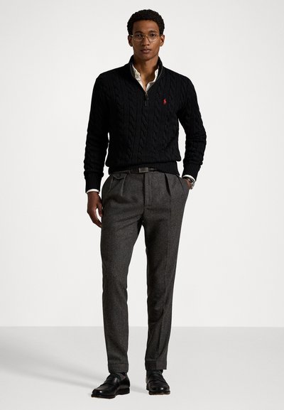 Maglione nero a costine con colletto alto, abbinato a pantaloni grigi a pieghe. Scarpe in pelle nere e un orologio d'argento completano il look.