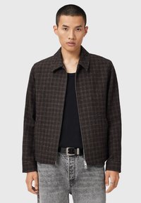 Veste zippée marron à carreaux avec un col pointu et une coupe décontractée, fabriquée dans un tissu texturé, associée à un jean en denim gris.