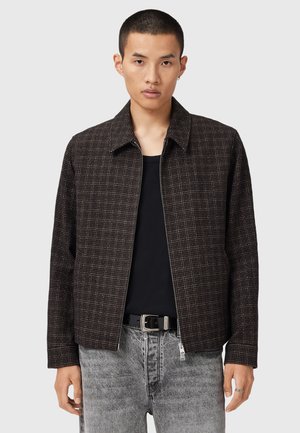 Veste zippée marron à carreaux avec un col pointu et une coupe décontractée, fabriquée dans un tissu texturé, associée à un jean en denim gris.