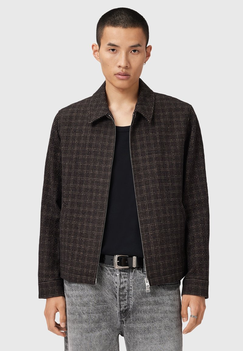 Brun ternet jakke med lynlås foran, spids krave og afslappet pasform, lavet af et tekstureret stof, parret med grå denim jeans.