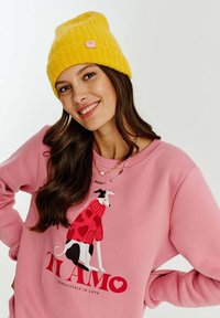 Růžová mikina s potiskem psa v červené a černé barvě, doplněná svetr srdíčkového vzoru. Celý outfit doplňuje jasně žlutá pletená čepice.