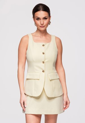 Femme aux cheveux foncés portant une robe crème sans manches avec des boutons dorés et des poches à rabat à l'avant, debout devant un fond clair uni.