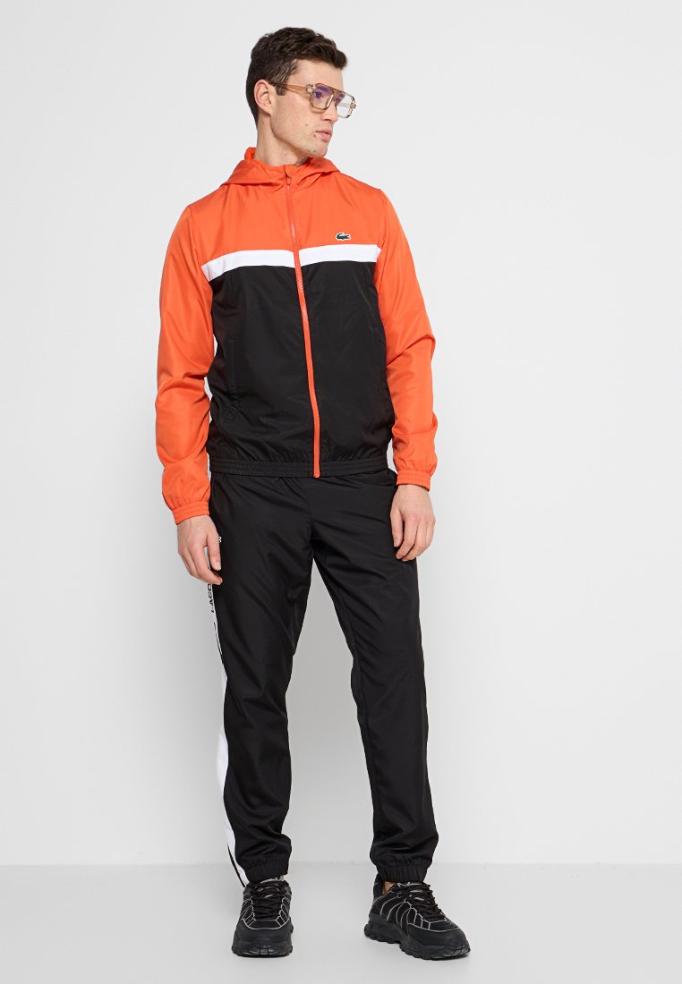 Veste coupe-vent orange et noire avec zip, bande blanche sur la poitrine, pantalon noir assorti et baskets noires texturées.