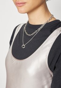 Femme portant des colliers en chaînes d'argent superposés sur une chemise noire et un top sans manches en satin beige clair.