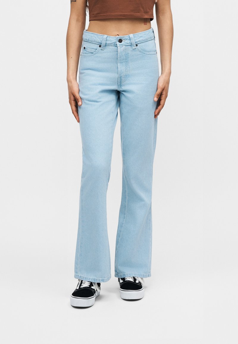 Hellblaue, tief sitzende Jeans, getragen von einer stehenden Person, kombiniert mit schwarz-weißen Sneakers und einem kurzen braunen Top.