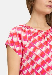 Kortærmet bluse i pink og rød med et geometrisk mønster. Har en samlet halslinje og en lille nøglehulldetalje foran.