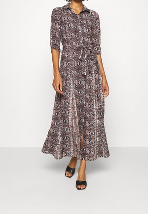 Frau trägt ein langärmliges, mit Gürtel versehenes Midikleid mit Schlangenprint, Kragen und vorderem Schlitz, kombiniert mit schwarzen, offenen Sandaletten mit Absatz.