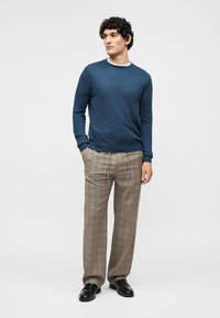 Άνδρας όρθιος με τα χέρια στις τσέπες φοράει μπλε πουκάμισο μακρυμάνικο, μπεζ καρό παντελόνι και μαύρα παπούτσια σε μονόχρωμο φόντο.