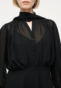 Femme portant un chemisier noir transparent avec un devant boutonné, des manches longues bouffantes et une écharpe noire unie drapée autour du cou.