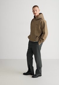 Sudadera con capucha marrón y bolsillo frontal, combinada con pantalones gris oscuro y botas negras. Textura suave y corte relajado.