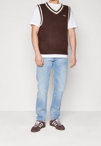 Chaleco de punto marrón con cuello en forma de V blanco, llevado sobre una camiseta blanca. Combinado con jeans azul claro y zapatos deportivos de color púrpura oscuro.