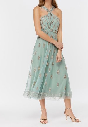 Een muntgroene jurk tot op de knie met een halterhals, versierd met gouden accenten en een vloeiende, doorschijnende laag aan de onderkant.