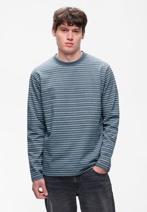 Jeune homme aux cheveux bruns portant une chemise à manches longues rayée bleue et grise et un jean foncé, debout devant un fond blanc uni.