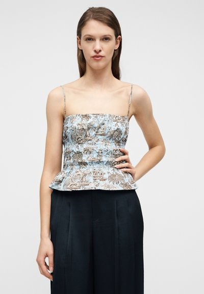 Γυναίκα που φοράει ένα ανοιχτό μπλε τοπ με λουλούδια και λεπτές τιράντες, καθώς και μαύρο, πλισέ παντελόνι με ψηλή μέση, στέκεται με το ένα χέρι στη μέση.