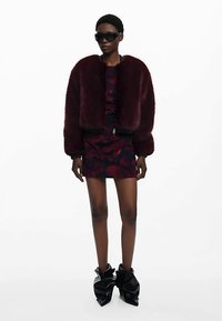 Viininpunainen tekoturkisbomber-takki tumman kukkakuvioisen minimekon päällä. Mallilla on mustat aurinkolasit ja veistokselliset mustat saappaat. Sileä pinta.