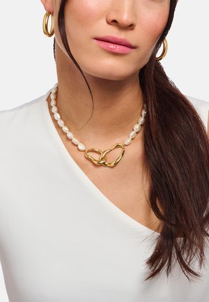 Frau mit langen braunen Haaren, die ein weißes Oberteil, goldene Creolen und eine Perlenkette mit zwei großen, verbundenen goldenen Herzanhängern trägt.