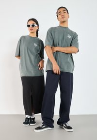 Grå oversized t-shirts, sort denim nederdel med split og løse marineblå jeans. Hvide sneakers med sorte accenter; solbriller fuldender looket.