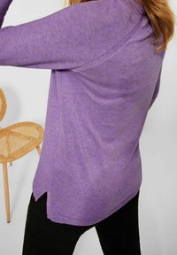 Maglione viola lavorato a maglia con maniche lunghe, caratterizzato da una vestibilità comoda, fessure laterali e una finitura texturizzata. Visibili pantaloni neri e una sedia di legno.