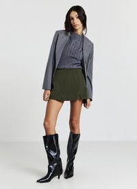 Blazer grigio sopra un maglione grigio testurizzato, abbinato a una gonna di velluto a coste verde. Stivali neri lucidi alti fino al ginocchio con punte appuntite.