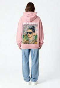 Felpa oversize rosa con stampa sul retro di una donna con occhiali da sole, elementi decorativi e la scritta "L'ARTE È COSTOSA" sopra.