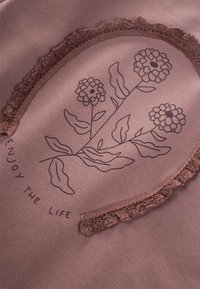 Dammrosafärgad tyg med oval design och svarta blommor. Texten lyder "NJUT AV LIVET." Accentuerad med en rynkad kant.